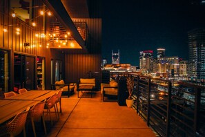 Terraza al aire libre con vistas a la ciudad, con una mesa y sillas de madera, luces colgantes y una barandilla.