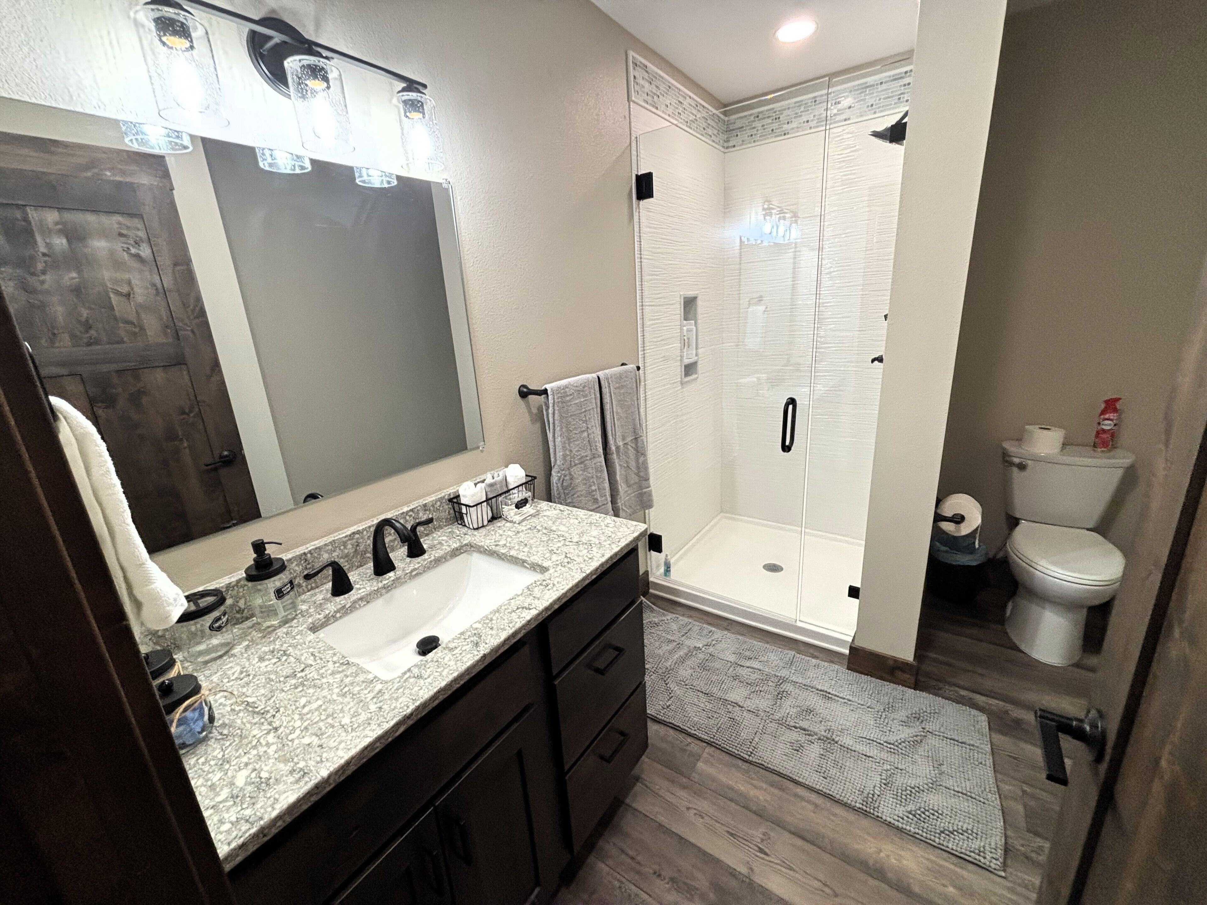 A modern bathroom with a granite countertop sink, a large mirror, a walk-in shower, and a toilet.