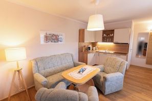 Una sala de estar con un sofá, un sillón y una mesita con una bandeja. Hay una lámpara, un cuadro en la pared y una zona de cocina al fondo.