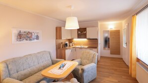Een woonkamer met een bank, fauteuil en een tafel, een keuken met houten kasten, en een gang die leidt naar een deur.