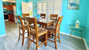 A dining area with a wooden table and chairs, a blue wall, and a kitchen in the background. - MANATEE COTTAGE:  Gulf Front 2-bedroom house (Hudson)
