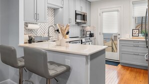 Une cuisine moderne avec des meubles gris, un îlot central doté de tabourets de bar, un four à micro-ondes et une grande fenêtre avec des stores.