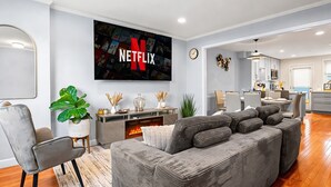 Un salon moderne avec un canapé gris, un téléviseur à écran plat affichant le logo de Netflix, un coin repas et une cuisine.