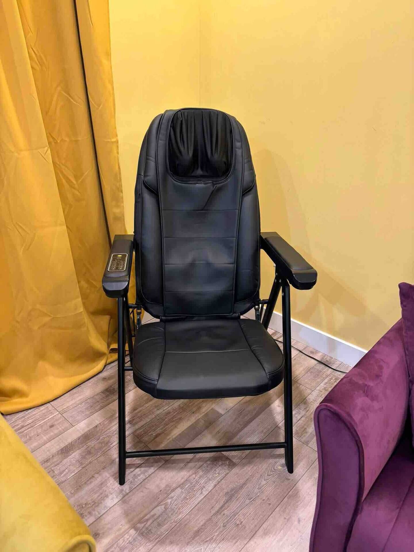A black office chair with armrests and a footrest, positioned against a yellow wall with a wooden floor.