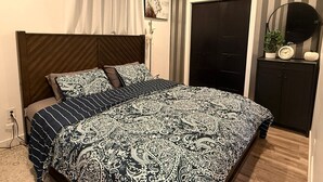 A bedroom with a bed, a dark headboard, a white curtain, a door, a small table with a clock, and a wall-mounted mirror. - HOME AWAY FROM HOME (Winnipeg)