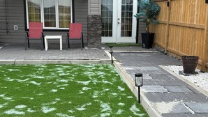 A backyard with artificial grass, a patio with chairs, a wooden fence, and a potted plant. - HOME AWAY FROM HOME (Winnipeg)