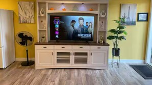 A living room with a flat-screen TV mounted on a white cabinet, a fan, and a potted plant. - HOME AWAY FROM HOME (Winnipeg)