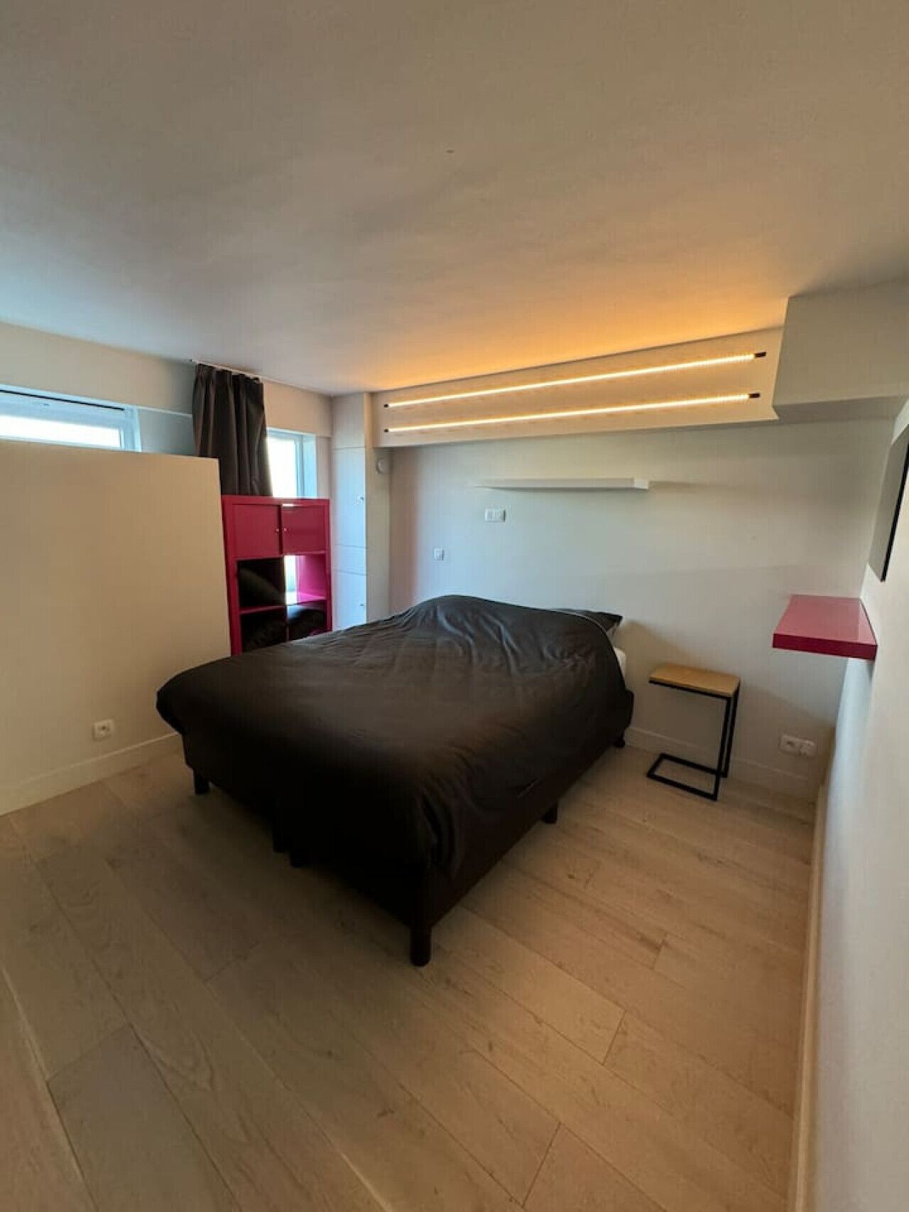 Une chambre à coucher avec un lit, une petite table de chevet, une étagère rouge et une fenêtre avec des rideaux.