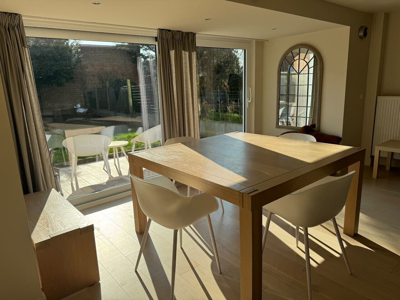 Un coin repas avec une table en bois et quatre chaises blanches, une porte vitrée donnant sur l’extérieur et une grande fenêtre en arc de cercle.
