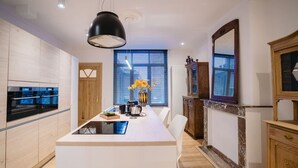 A modern kitchen with a central island, built-in appliances, and a large window with blinds. - Circa 1929 - Suite met keuken (Ostend)