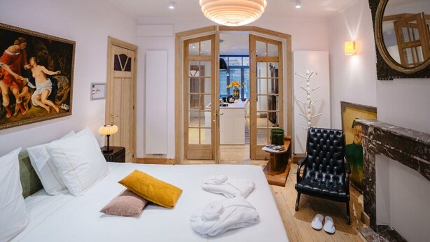 A hotel room with a bed, a painting, a wooden door, a black chair, and a fireplace. - Circa 1929 - Suite met keuken (Ostend)
