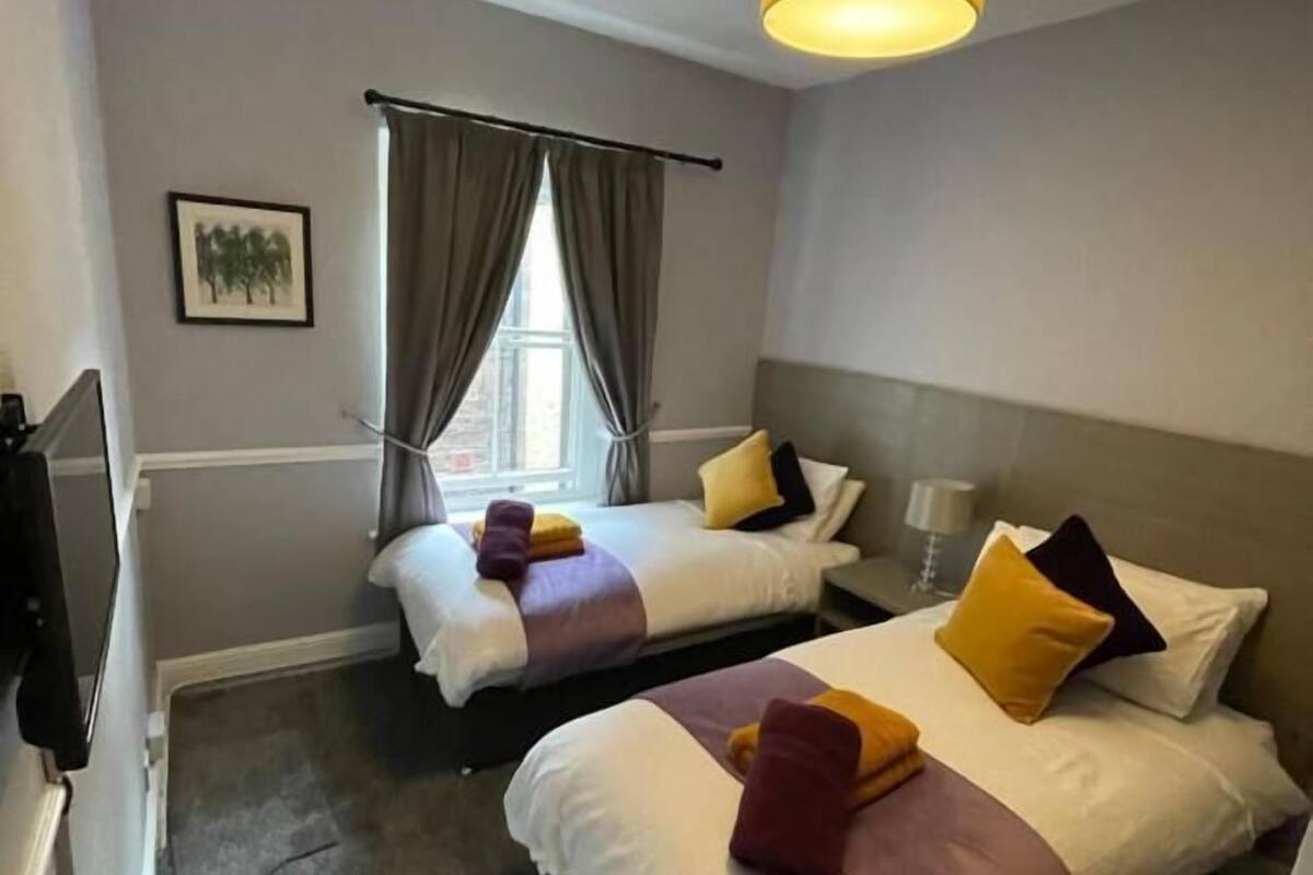 Quarto de hotel com duas camas, uma janela com cortinas, uma luminária no teto e um quadro pendurado na parede.