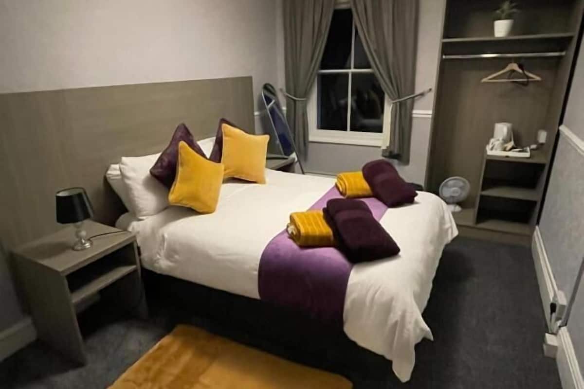 Quarto de hotel com cama, mesa de cabeceira, abajur e um tapete pequeno.