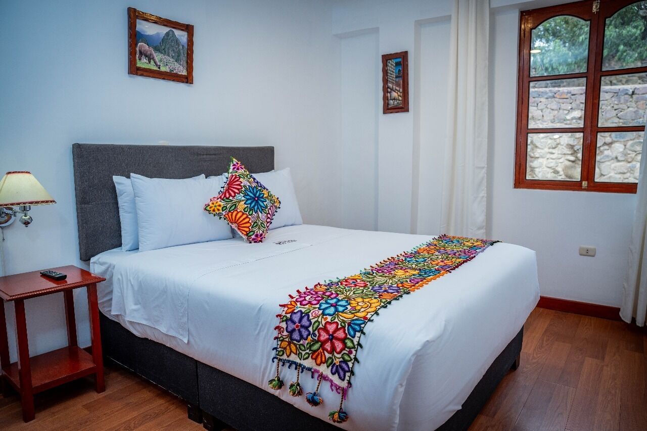 Una cama bien hecha con un colorido edredón bordado, una mesita de noche con una lámpara y cuadros enmarcados en las paredes.
