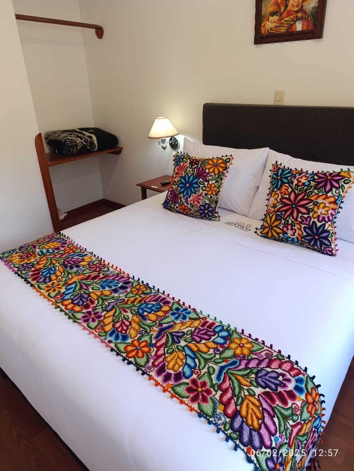 A bed with a colorful embroidered bedspread, two pillows with floral patterns, a bedside lamp, and a framed picture on the wall.