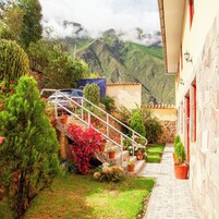 Un patio con una escalera de piedra que conduce a un edificio, rodeado de macetas con plantas y un coche aparcado cerca.