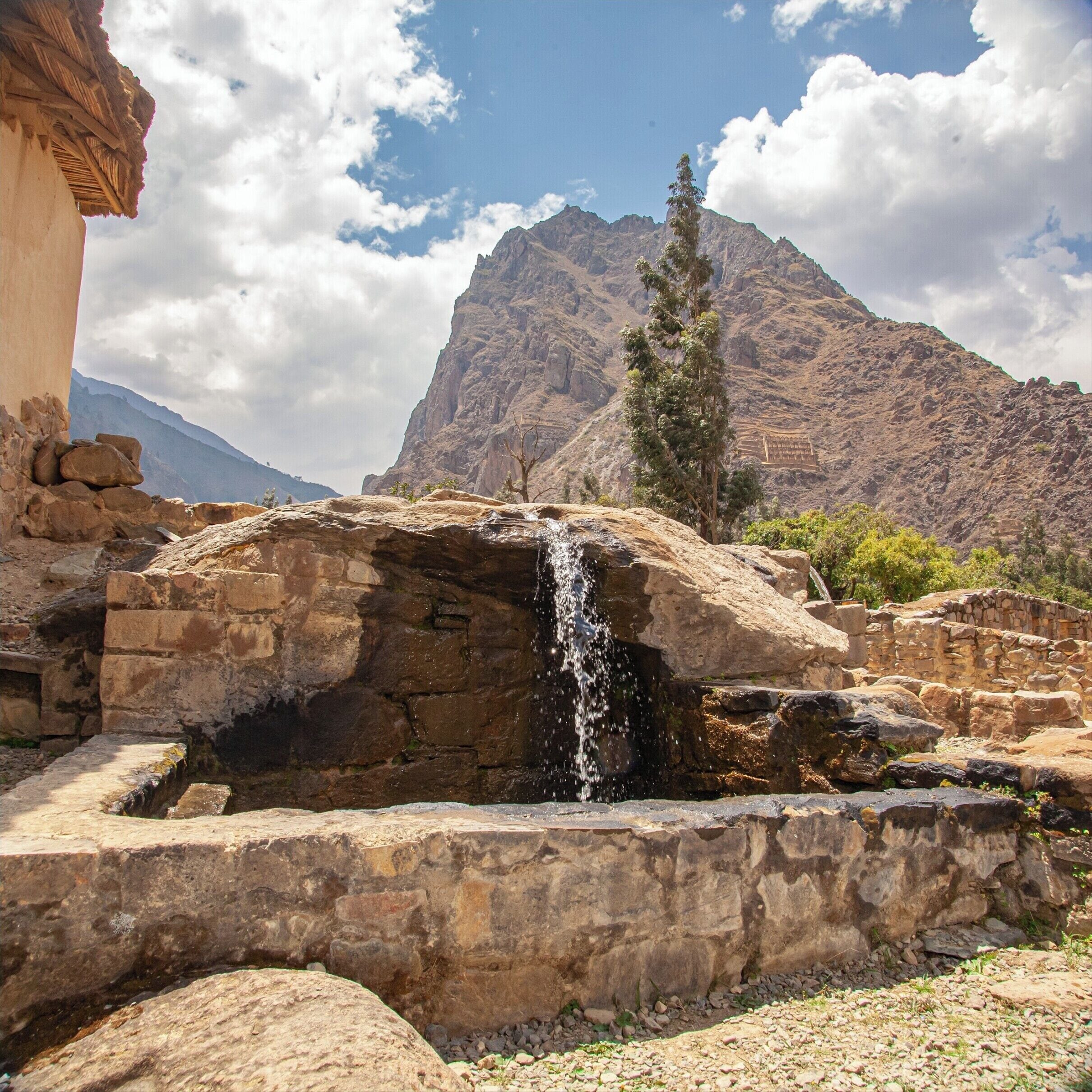 Una estructura de piedra con una cascada, rodeada de montañas y un cielo despejado.
