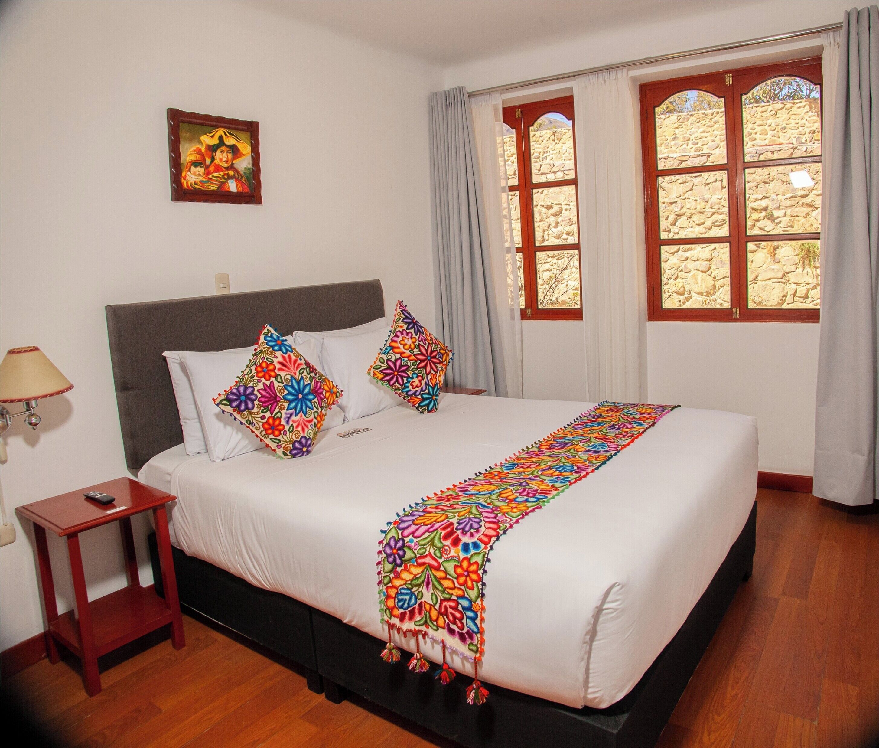 A neatly made bed with white linens and colorful floral pillows, a wooden nightstand with a lamp, and a stone wall visible through a window with curtains.
