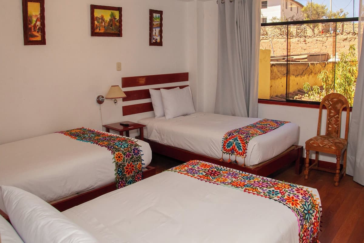 Een hotelkamer met drie eenpersoonsbedden, een houten hoofdbord, een stoel en een raam met uitzicht op een gebouw en bomen.