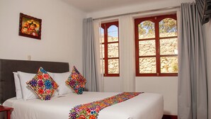 Een net opgemaakt bed met een kleurrijke geborduurde deken, een houten hoofdbord en een stenen muur zichtbaar door een raam met gordijnen.