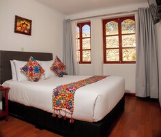Una cama bien hecha con un colorido edredón bordado, un cabecero de madera y una pared de piedra visible a través de una ventana con cortinas.