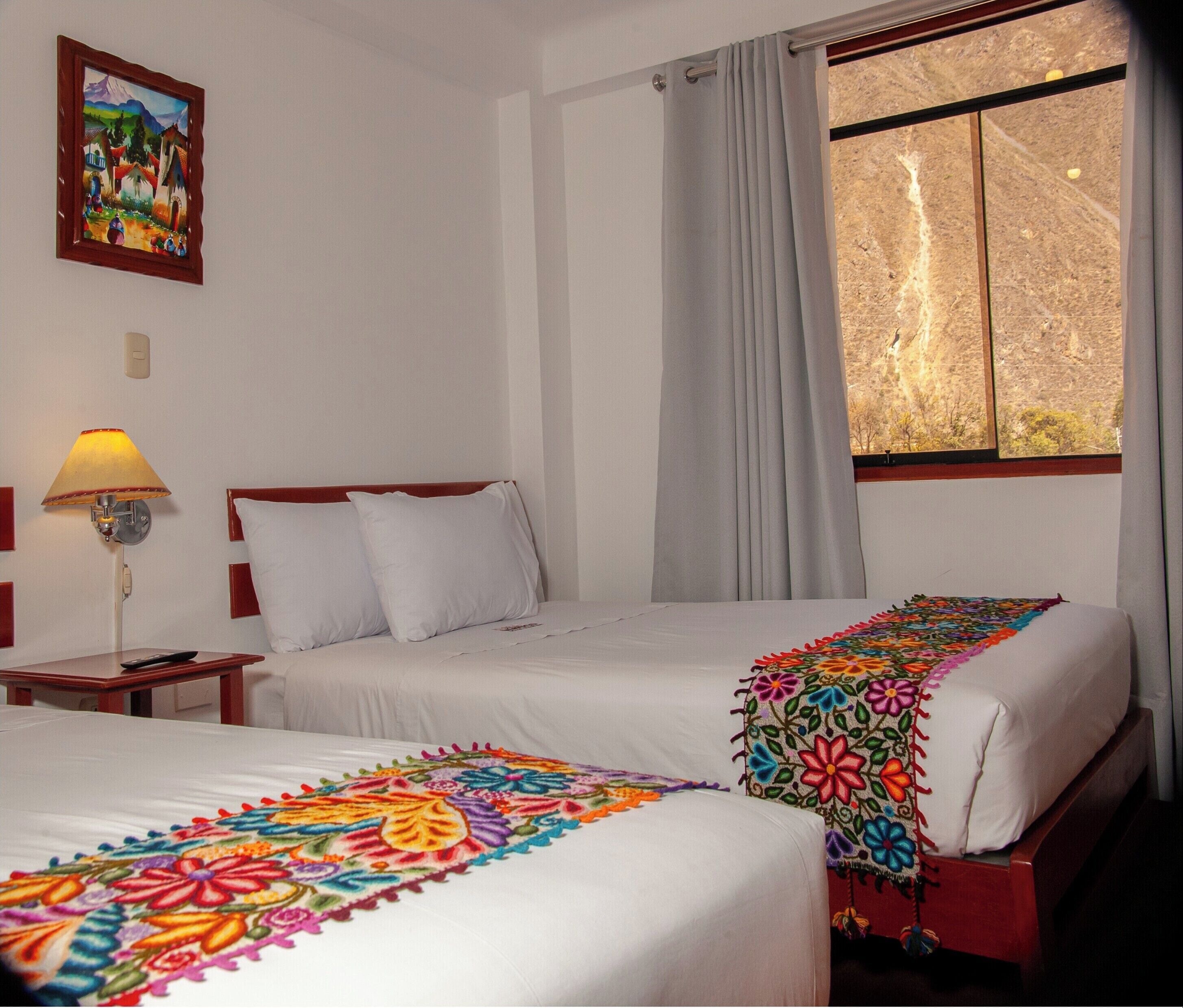 Habitación de hotel con dos camas, una mesita de noche con lámpara, un cuadro en la pared y una ventana con cortinas.