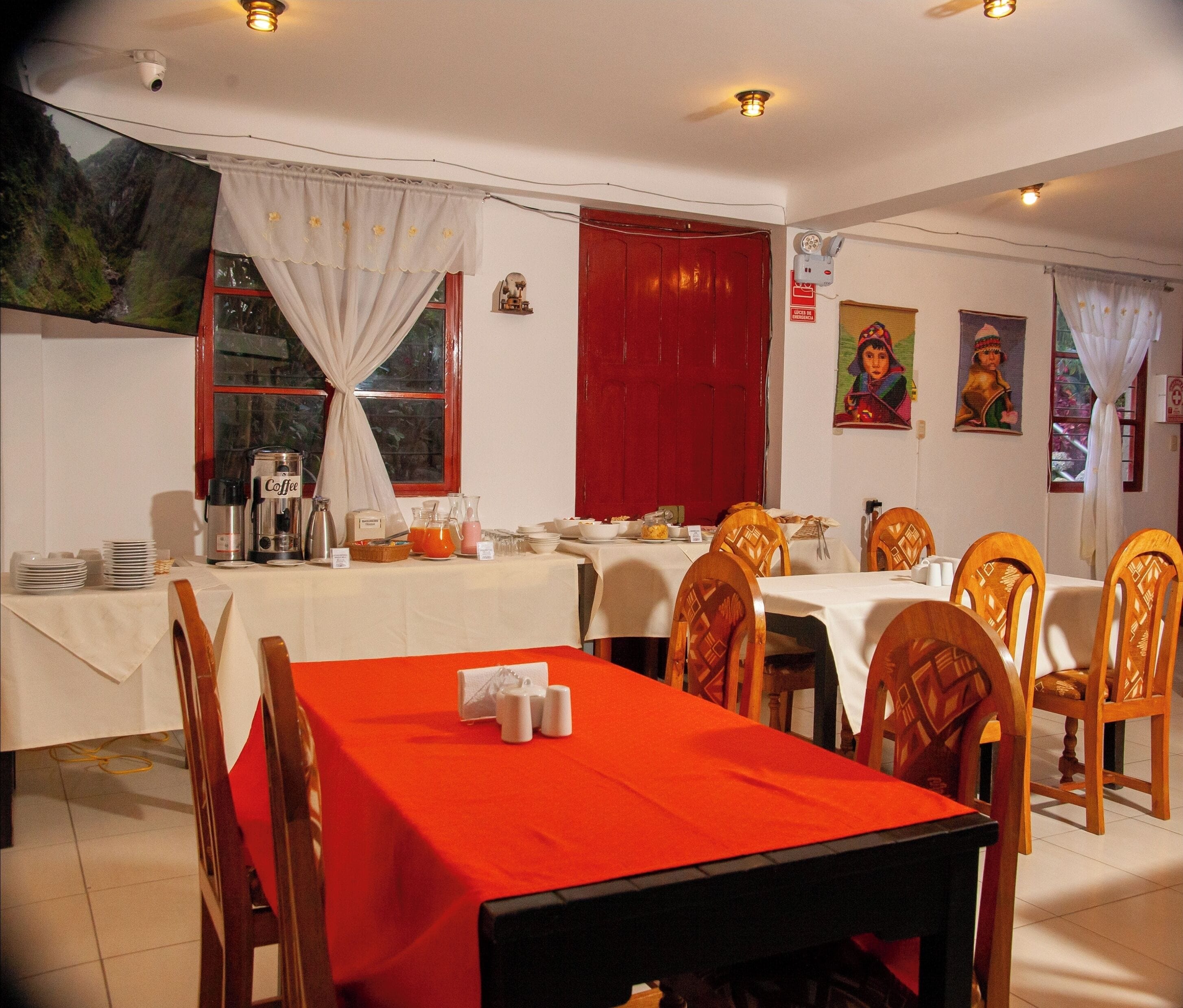 Un comedor con mantel rojo, mesas cubiertas con mantel blanco y sillas de madera.