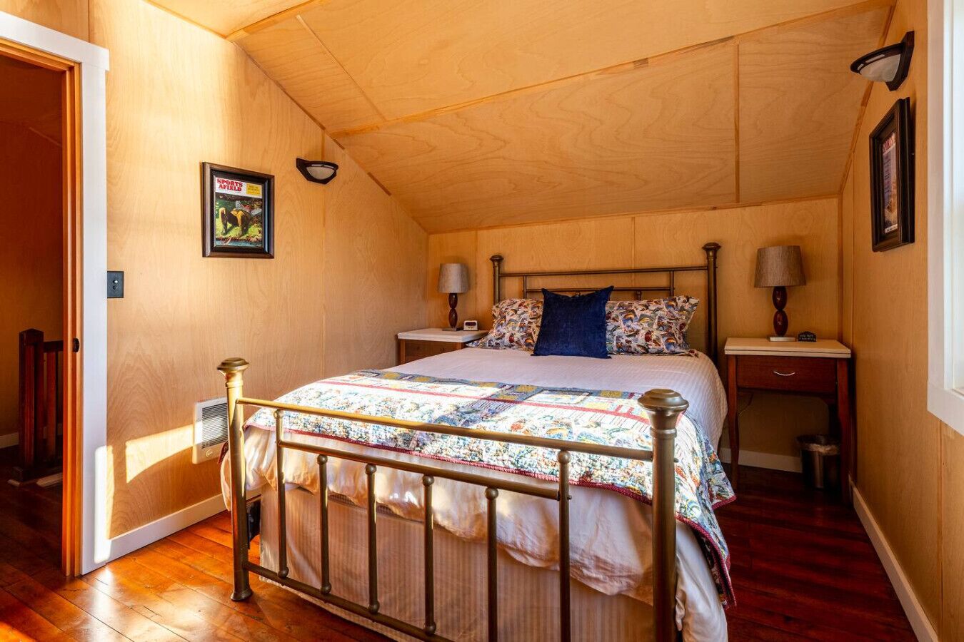 A bedroom with a wooden ceiling, a metal bed frame, a quilted bedspread, two bedside tables with lamps, and a framed picture on the wall.