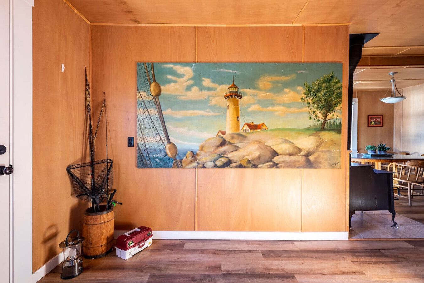 A room with a wooden floor, a painting of a lighthouse, a fishing net, and a lantern.