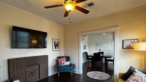 Una sala de estar con chimenea, un sillón azul, una mesa de comedor con sillas, un sofá amarillo y un ventilador de techo.
