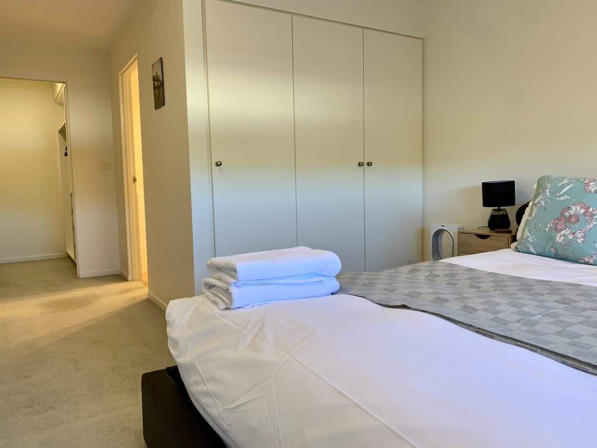 Una habitación de hotel con una cama, un armario, una mesita de noche con una lámpara y un cojín estampado con motivos florales.