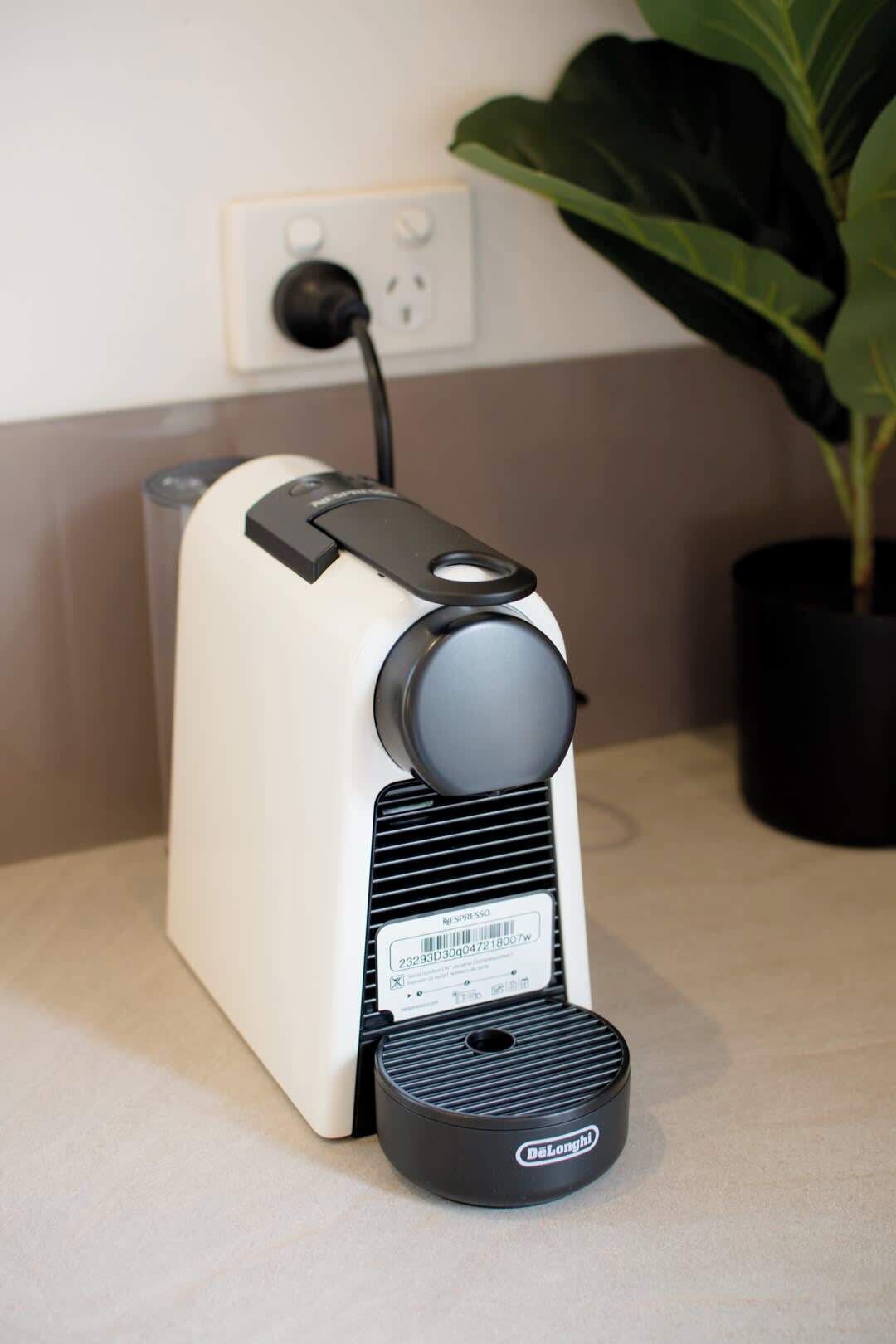 Una cafetera Nespresso con un portacapsulas de la marca Delonghi, conectada a un enchufe, y una planta en maceta al fondo.