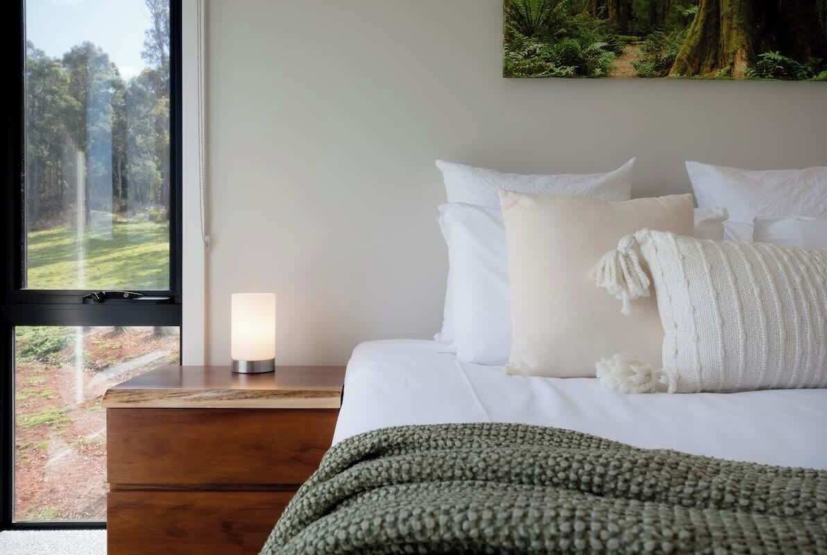A bedroom with a bed, pillows, a bedside table with a lamp, a window with a view of a forest, and a painting on the wall.