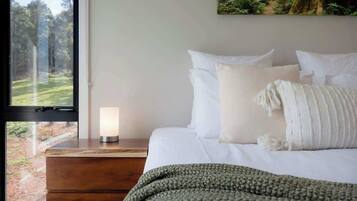 A bedroom with a bed, pillows, a bedside table with a lamp, a window with a view of a forest, and a painting on the wall.