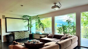A modern living room with a large sectional sofa, a flat-screen TV, and a large window offering a view of greenery and a balcony. - Charming bungalow house in Sherman Oaks w/ pool table, fire pit, hot tub & piano (Los Angeles)