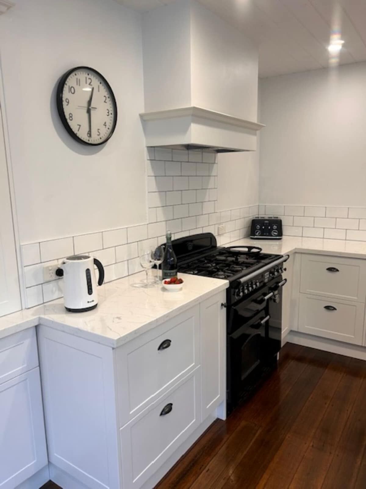 Une cuisine avec des meubles blancs, un poêle noir, une bouilloire et une horloge au mur.