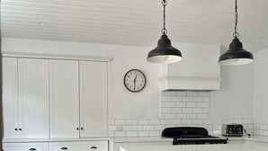 Une cuisine avec des meubles blancs, un îlot central noir doté de tabourets, une horloge au mur et des luminaires suspendus.