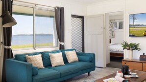 A teal sofa with patterned cushions, a coffee maker, and a view of a lake through a window with curtains. - Waterfront Cottage - direct waterfront access, boat-friendly block, firepit, water views (Swan Bay)