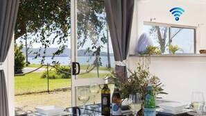 A table with wine bottles and glasses, a view of a lake, and a window with a curtain. - Waterfront Cottage - direct waterfront access, boat-friendly block, firepit, water views (Swan Bay)