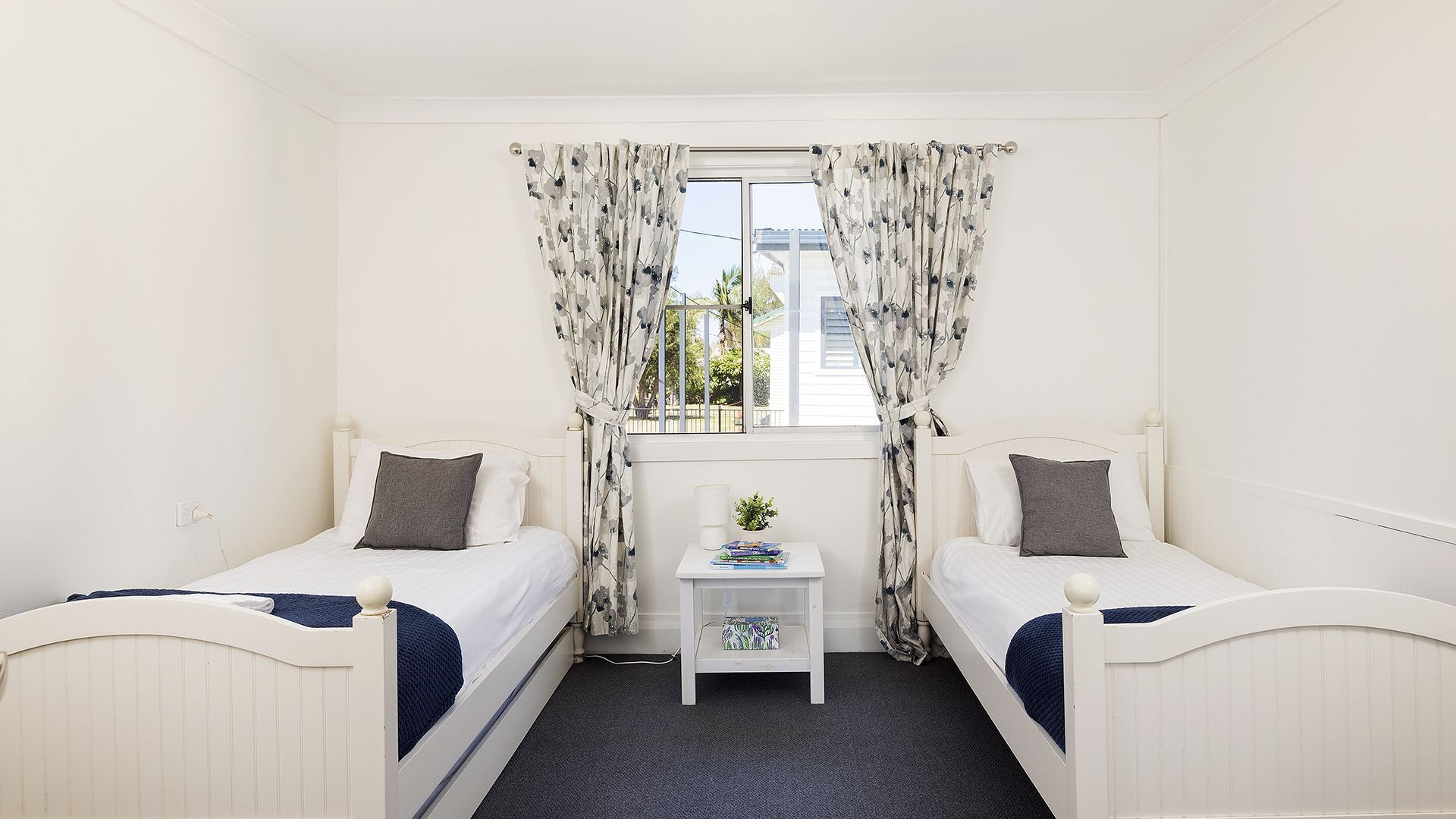 A room with two single beds, a small bedside table with a plant, and a window with floral curtains.