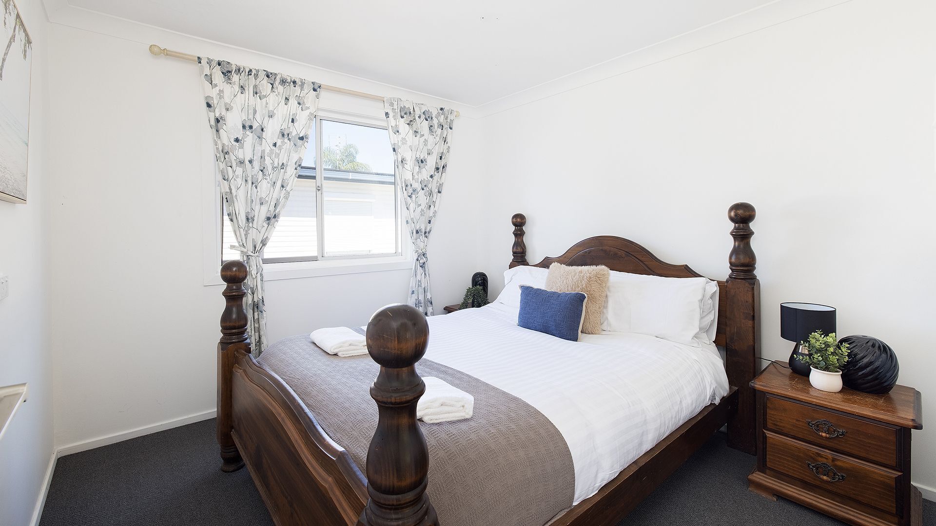 A bedroom with a wooden four-poster bed, a nightstand with a lamp, and a window with floral curtains.