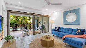 A modern living room with a blue sofa, a coffee table, and a large window overlooking a garden. - Temple Breeze - Swim Out Apartment (Port Douglas)