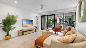 Una sala de estar moderna con sofá, mesa de centro y televisor. Cuenta con un balcón con plantas y vistas al exterior.