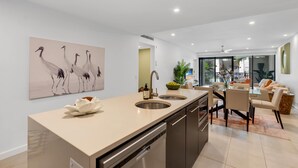 Una cocina moderna con una isla central, un comedor con mesa de vidrio y una sala de estar con sofá y planta.