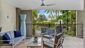 Ein Balkon mit Deckenventilator, einer Couch, einem Couchtisch und Stühlen. Im Hintergrund ist ein Pool und Palmen zu sehen.
