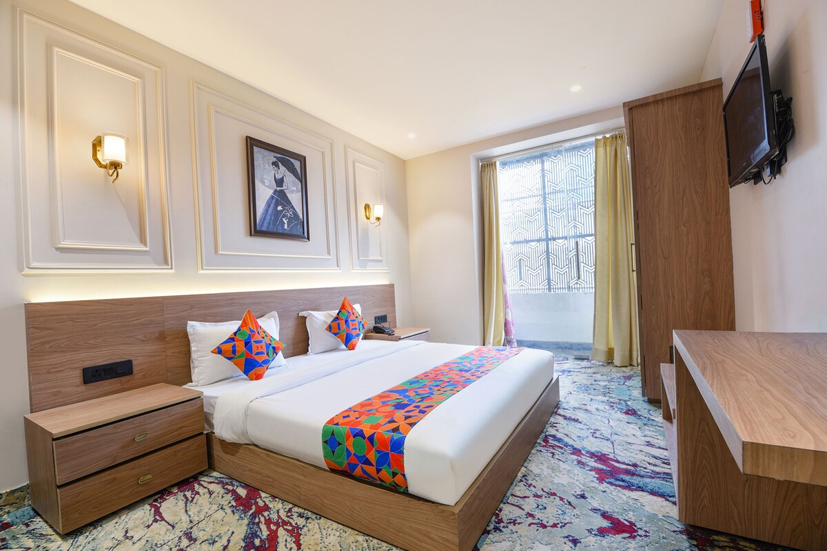 A hotel room with a large bed, a nightstand, a wooden desk, a TV mounted on the wall, and a window with curtains.