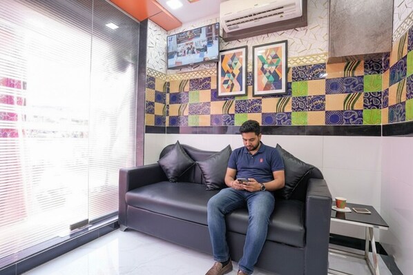 A man sitting on a dark leather couch in a modern room with patterned tile walls and a television mounted above. - Fabexpress Raj Palace (Mumbai)