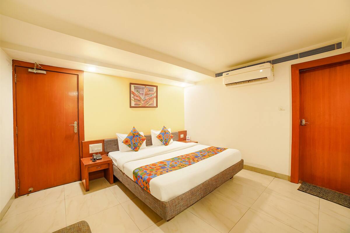 A hotel room with a large bed, wooden doors, an air conditioning unit, and a small bedside table.