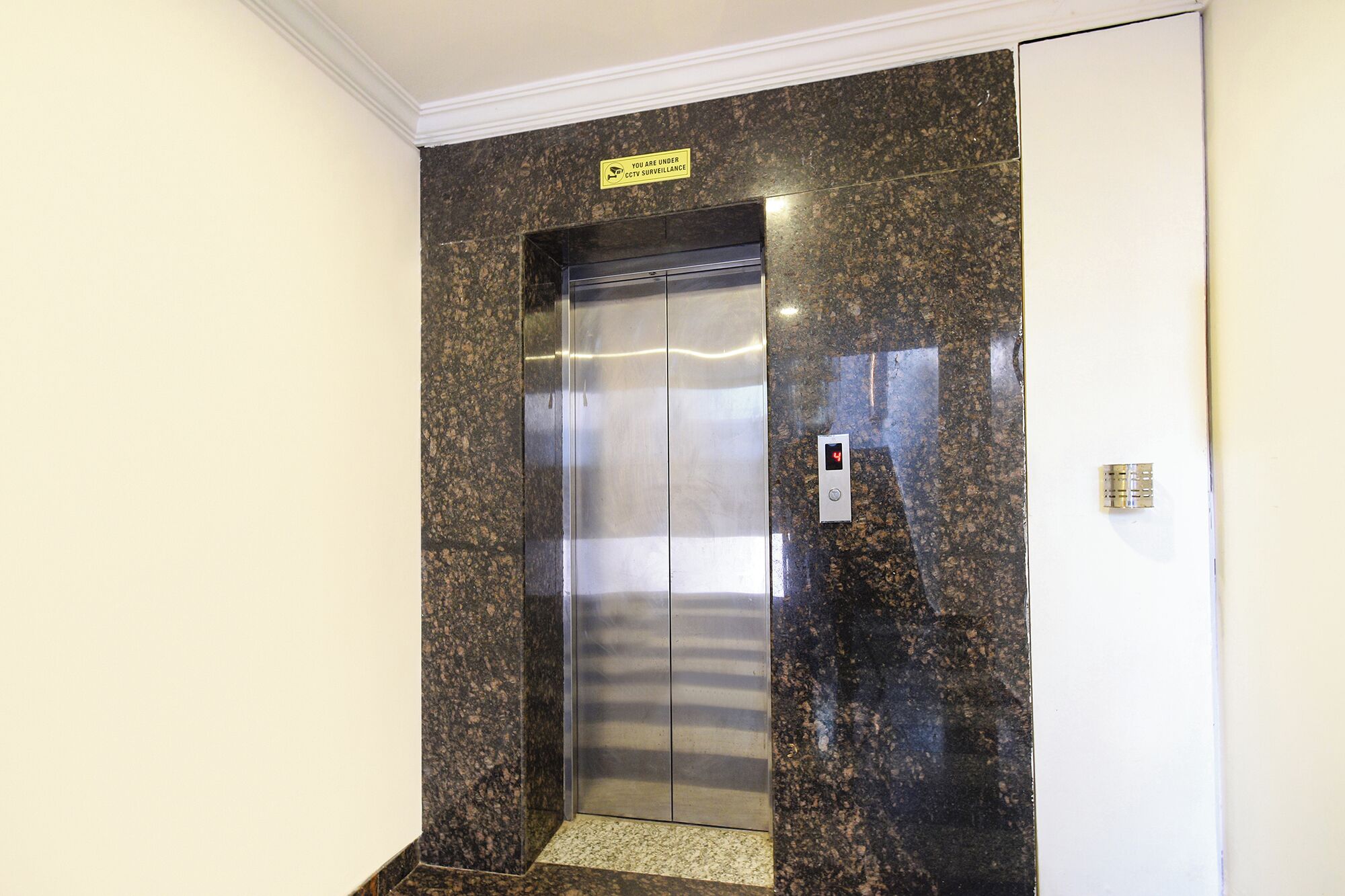 Un ascensor con una pantalla digital que muestra el número 4, un cartel de videovigilancia y una puerta metálica.