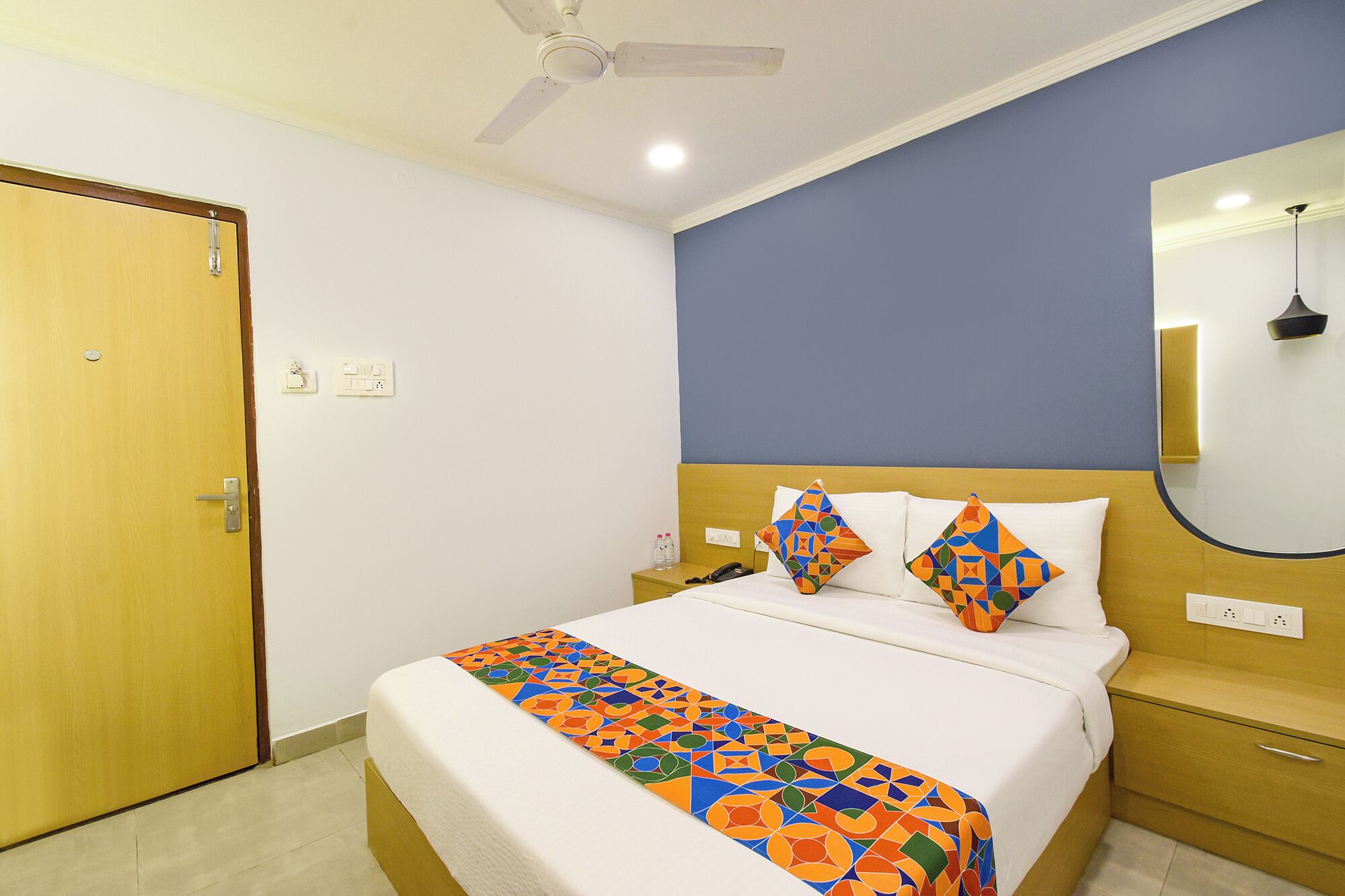 Une chambre d’hôtel avec un lit, une table de chevet, un miroir, un ventilateur de plafond et une porte.